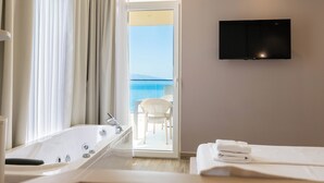 A hotel room with a bathtub, a TV mounted on the wall, a balcony with a chair, and a view of the sea.