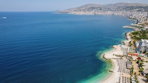 An aerial view of a coastal city with a beach, buildings, and a mountainous backdrop. - Mirage Bay Deluxe Saranda (Sarandë)