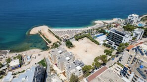 An aerial view of a coastal area with a beach, a parking lot, and several buildings including a hotel with a pool and a rooftop garden. - Mirage Bay Deluxe Saranda (Sarandë)