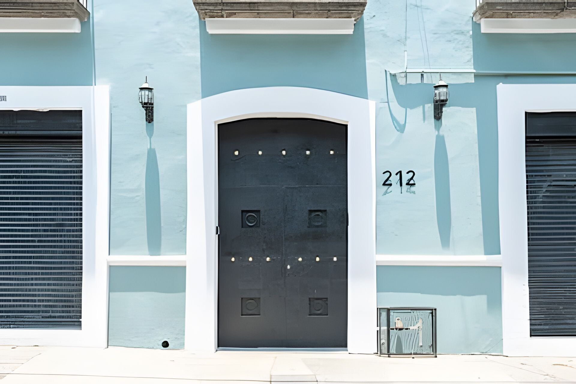 A building with a large, dark-colored door marked with the number 212, flanked by two closed shutters and two wall-mounted lights.