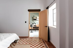 A bedroom with a bed, a checkered floor, a door leading to a kitchen with a window.