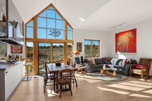 Un salón espacioso con una gran mesa de comedor, un sofá y una mesa de centro. Amplias ventanas ofrecen vistas al exterior.