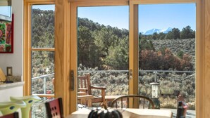 A dining area with a table and chairs, a wooden table, and a view of a forested landscape through large windows. - 25 Acre Artist Ranch, hot tub & tennis, near Aspen (Carbondale)