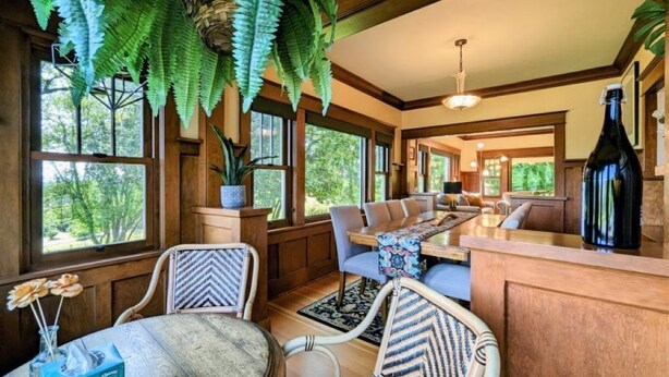 Un interior acogedor con una mesa de comedor de madera, sillas, un sofá y helechos colgantes.