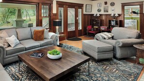 Un salón amplio con un sofá gris, un sillón gris y una mesa de centro de madera. Se observa una estantería con libros y adornos, y un ventanal que ofrece vistas al exterior.