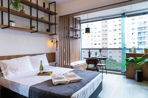 Una habitación de hotel moderna con una cama grande, mesita de noche y un balcón con vistas a la ciudad.