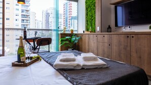Una habitación de hotel moderna con cama, televisor, aparador de madera, pared verde y balcón con mesa y sillas.