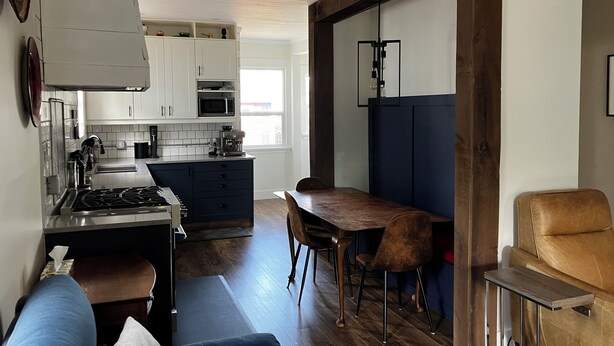 Una cocina moderna con armarios blancos, un microondas integrado y una cafetera. Hay un comedor con mesa y sillas de madera, y un sofá en la sala de estar.