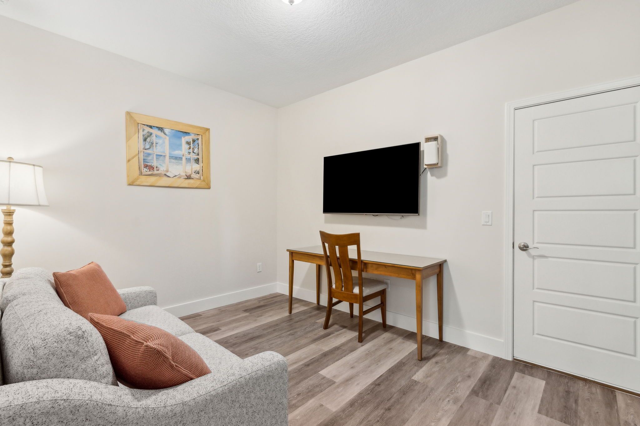 Una habitación con un sofá gris, un escritorio de madera, un televisor montado en la pared y una puerta blanca.
