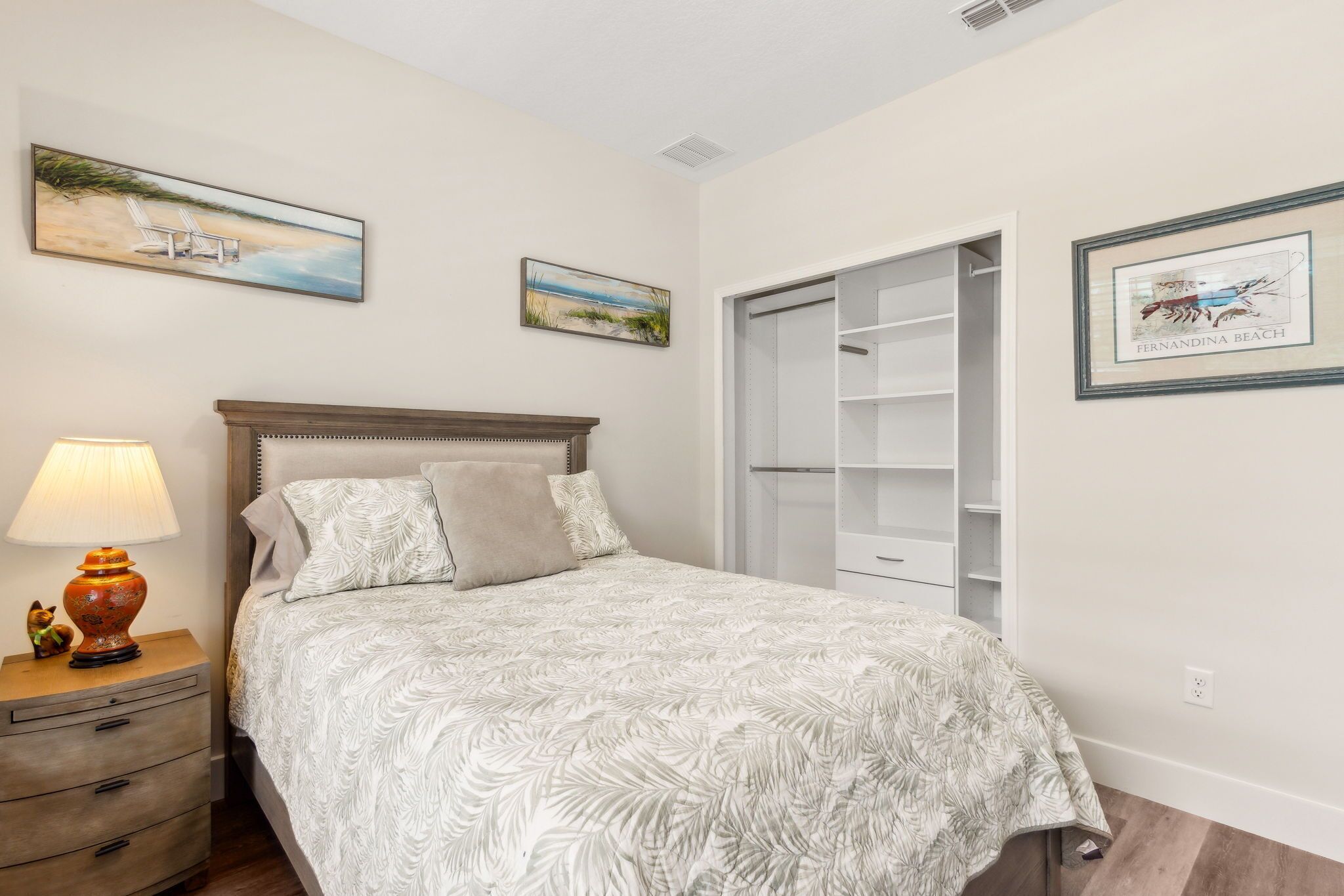 Quarto com cama, mesa de cabeceira e abajur, e guarda-roupa com portas abertas.
