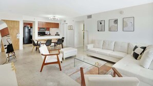 A modern living room with a white sofa, a glass coffee table, and a dining area with a wooden table and chairs. - Newly Renovated 3BR Condo with Bay Views & Parking (Miami)