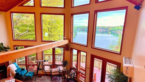 A spacious room with large windows offering views of a lake and trees. There is a wooden deck outside, a sofa, and a dining table with chairs inside. - Lake Cabin 8 acres private dock fire pit hot tub game room (Gouverneur)