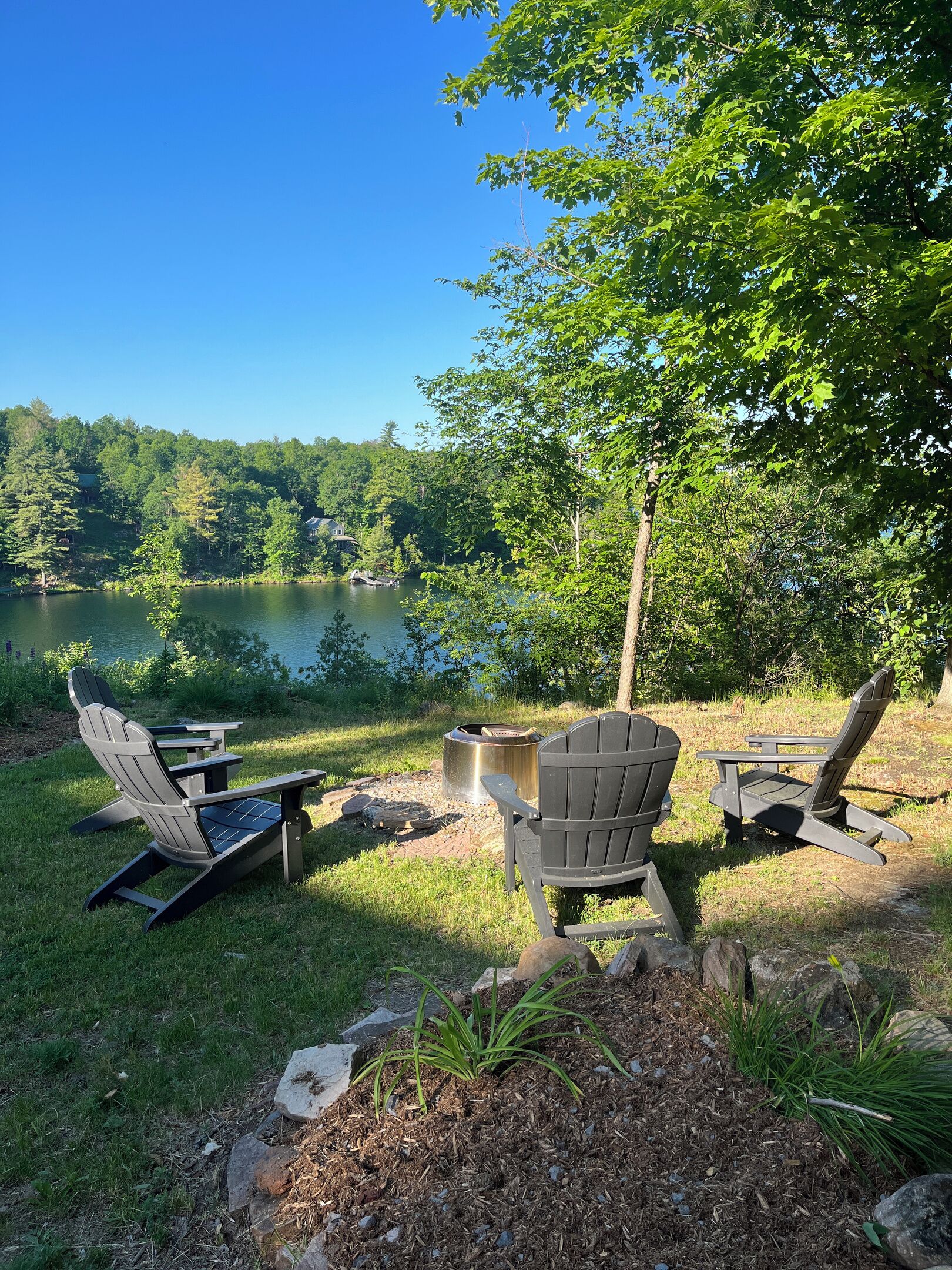 A lakeside setting with Adirondack chairs, a fire pit, and a view of a lake surrounded by trees.