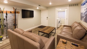 A living room with two leather sofas, a coffee table, a TV mounted on the wall, skis and ski equipment against the wall, and a mountain painting on the wall. - Location, location!  Spacious 4-BR double apartment near Cottonwood Canyons (Cottonwood Heights)