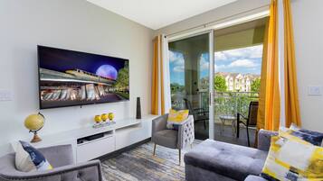 A modern living room with a flat-screen TV, a grey sofa, and a glass door leading to a balcony with yellow curtains.