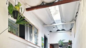 Un estrecho pasillo exterior con suelo de baldosas, una silla plegable negra y plantas en macetas.