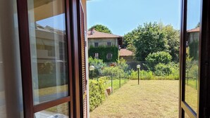 On aperçoit, à travers une porte vitrée, une cour avec une zone délimitée par une clôture, un sol carrelé et, au fond, un bâtiment dont le toit est recouvert de tuiles rouges.