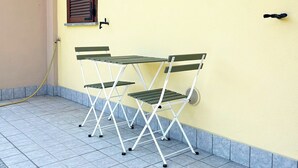 Un petit espace extérieur aménagé avec une table et des chaises, adossé à un mur jaune, une fenêtre avec des volets et un tuyau d’arrosage posé sur le sol.