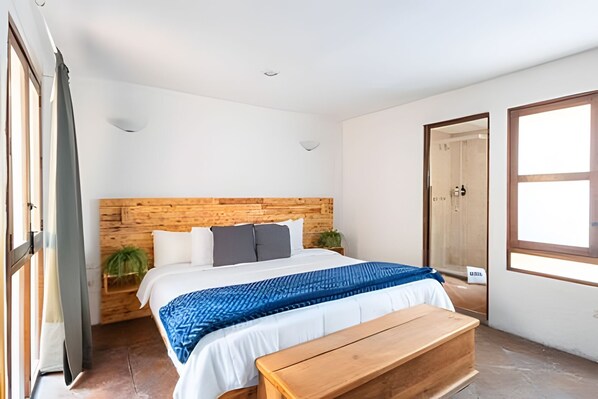 Un dormitorio con cabecera de madera, cama con ropa de cama blanca y azul, banco de madera y una planta en maceta.