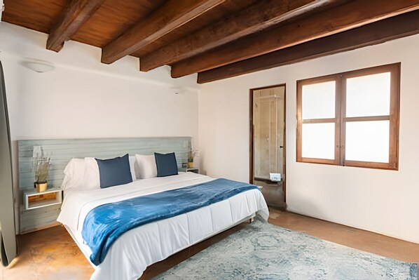 A bedroom with a bed, wooden ceiling beams, a large window, and a nightstand with a lamp.