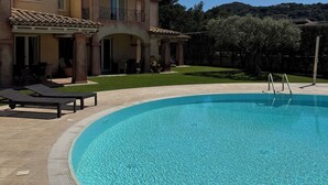 A swimming pool with a tiled deck, lounge chairs, and a building with a tiled roof in the background.