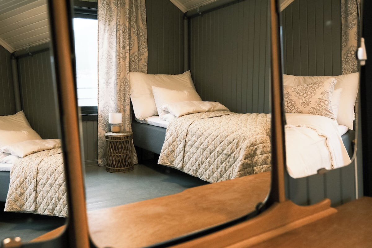 A mirrored view of a bedroom with a bed, pillows, a bedside table, and a lamp.