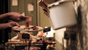 Una persona sostiene un plato con comida, un letrero que indica 'Brimi Speisekline (Brimi Sate)' y un recipiente metálico al fondo.