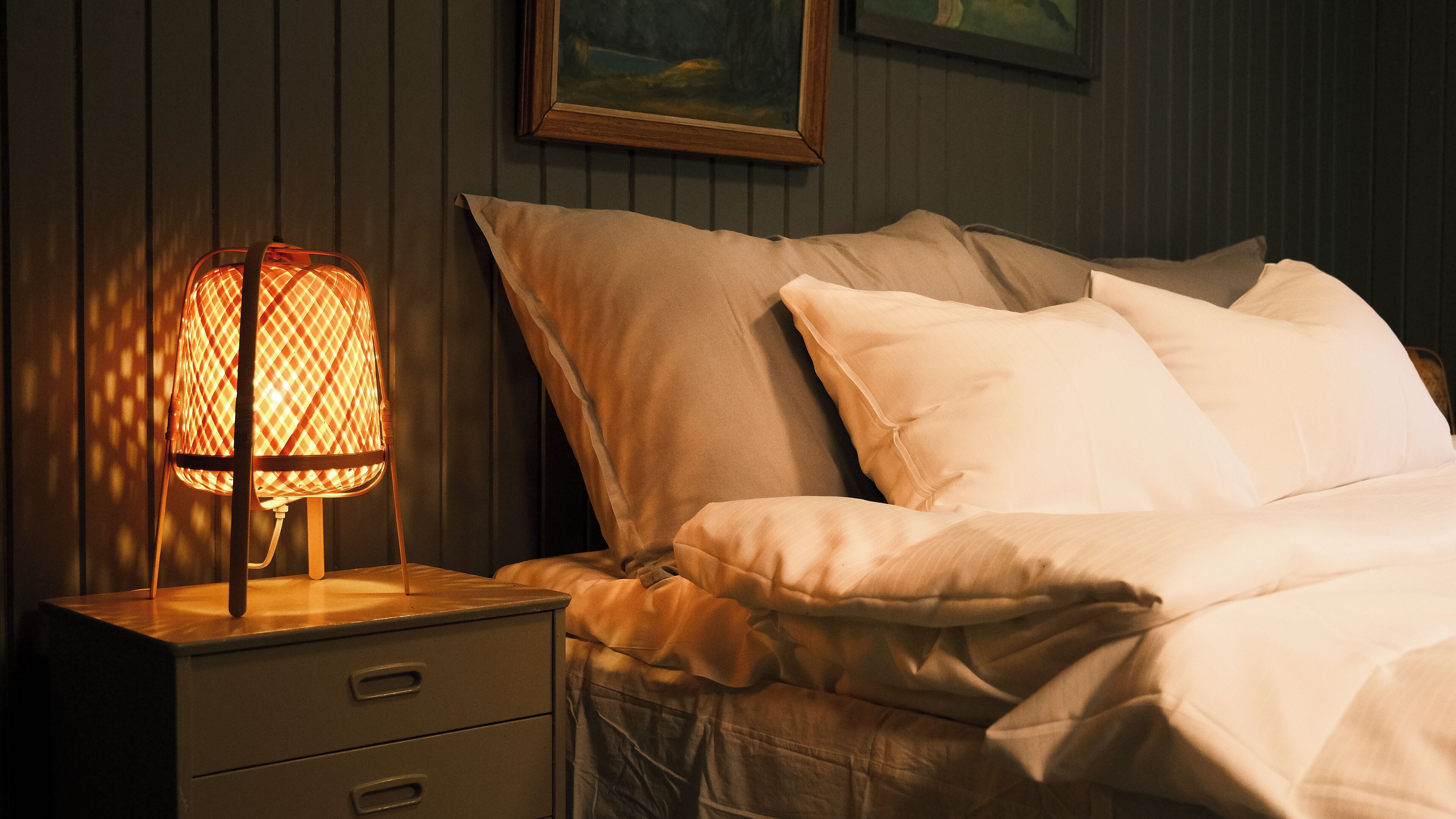 Una cama con ropa de cama blanca, una mesita de noche con lámpara y un cuadro en la pared.