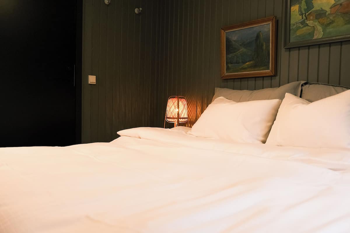 A neatly made bed with white linens, a wall-mounted lamp, and two framed artworks on the wall.