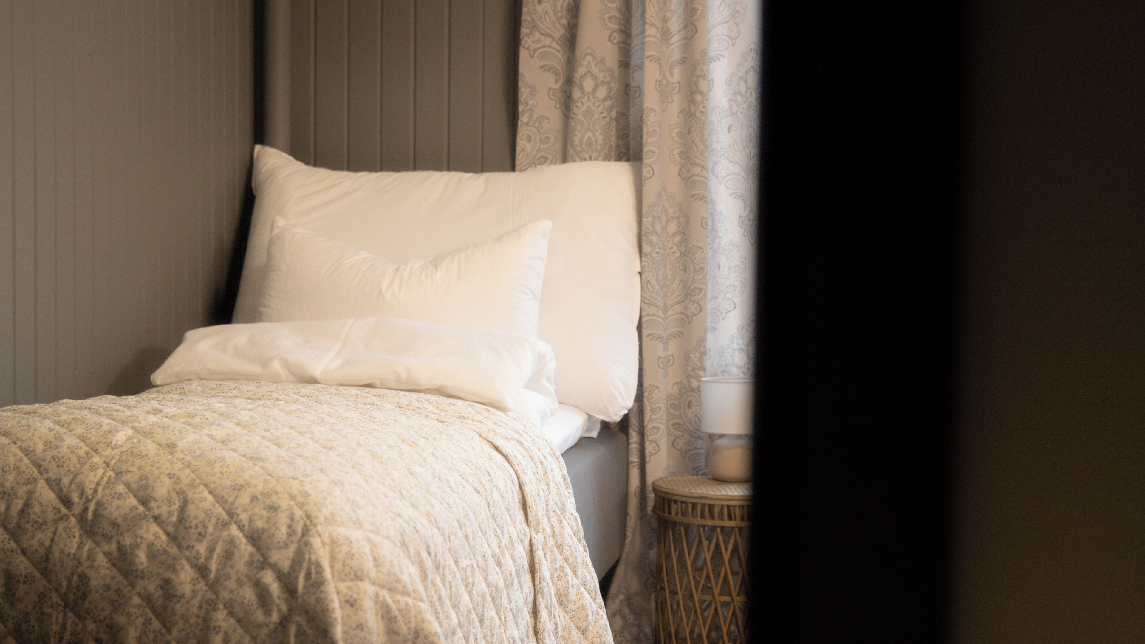 A bed with a quilted cover, a pillow, and a bedside table with a lamp.