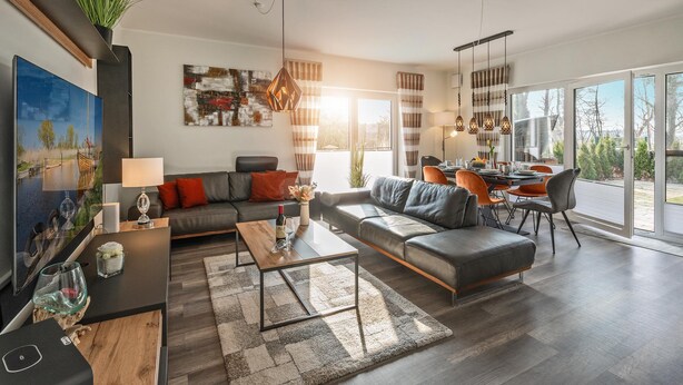 A modern living room with a sofa, coffee table, and dining area. There is a large window providing natural light and a view of the outdoors. - 5 star apartment for 6 persons, ground floor with fenced garden (Zinnowitz)