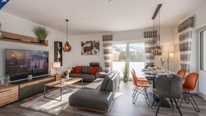 A modern living room with a flat-screen TV, a sofa, a dining table with chairs, and a large window with curtains. - 5 star apartment for 6 persons, ground floor with fenced garden (Zinnowitz)