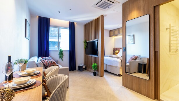 A modern hotel room with a wooden floor, a sofa, a dining table set for two, a TV, and a large mirror. - Próximo a Paulista com piscina  (Sao Paulo)
