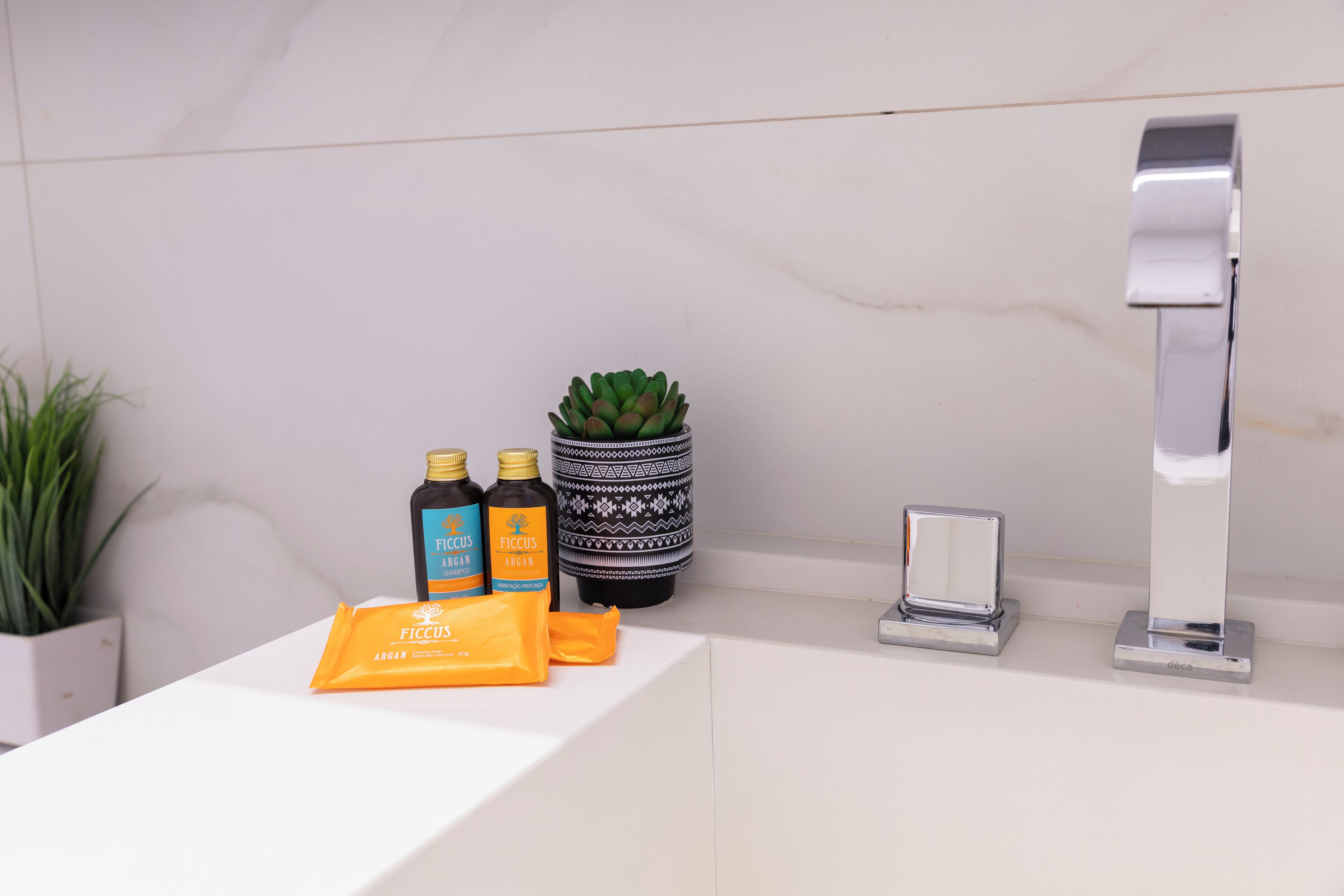A bathroom counter with a white sink, two bottles of Ficus Vegan body wash, a potted plant, and a mirror.