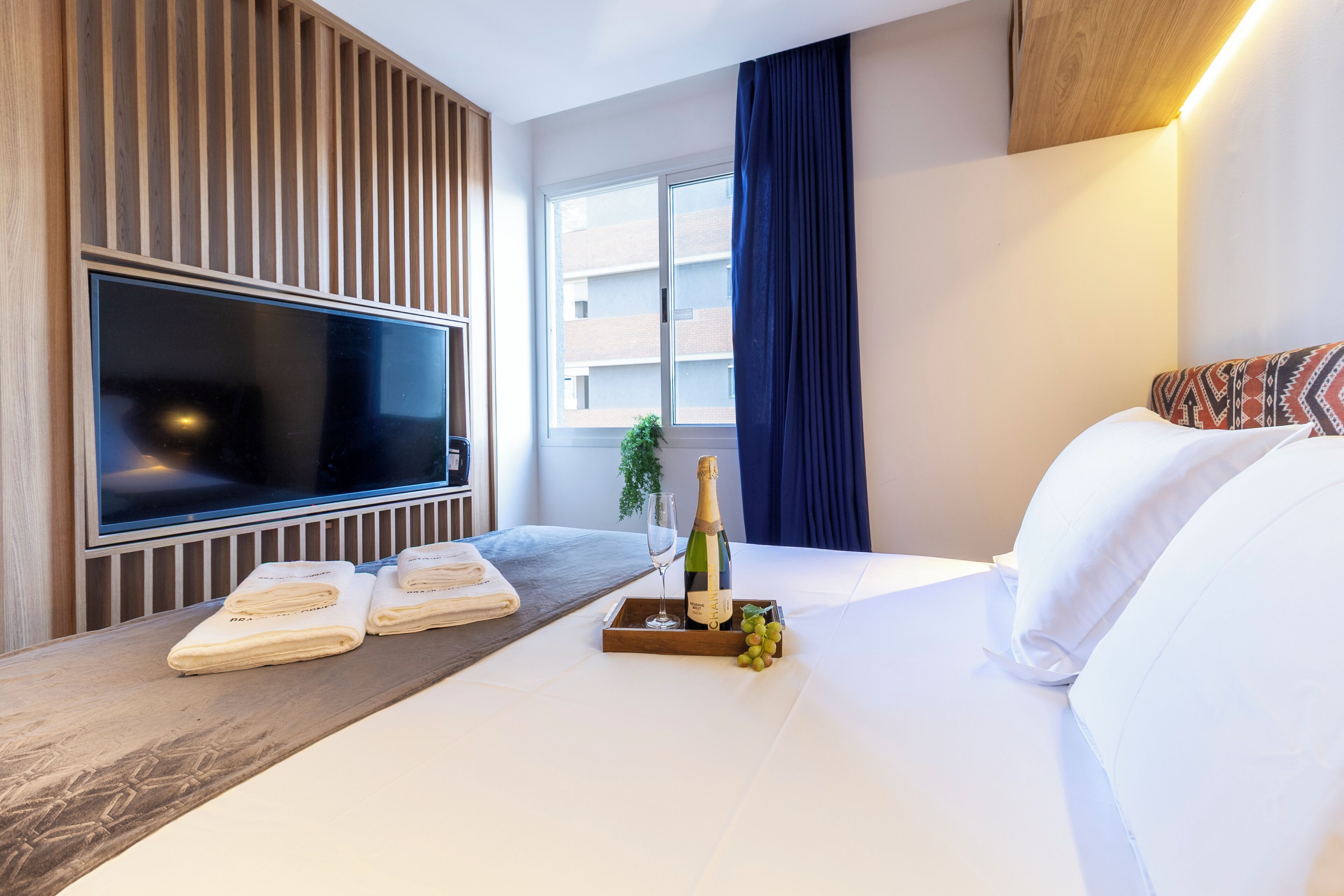Hotellihuone, jossa on suuri litteänäyttöinen televisio, puinen sivupöytä, jossa on pyyhkeitä, pullo samppanjaa ja tarjotinalus rypäleitä.