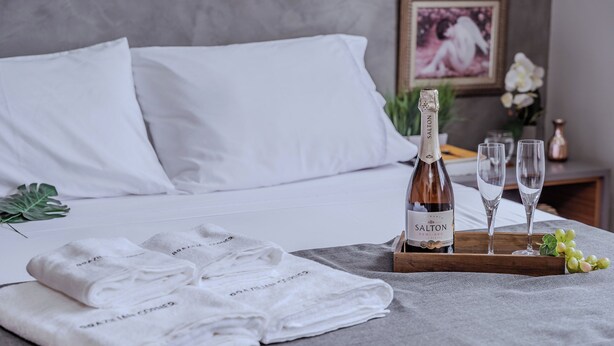 A neatly made bed with white linens, a bottle of Salton champagne, two glasses, and a tray with grapes. - 3 Quartos com vaga no Prado (Belo Horizonte)