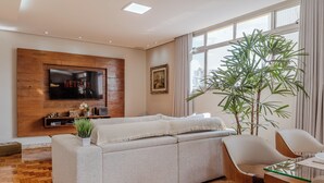 A modern living room with a sofa, a TV mounted on a wooden wall, a plant, and a dining area with chairs. - 3 Quartos com vaga no Prado (Belo Horizonte)