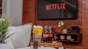 A cozy living room with a large screen displaying "NETFLIX," popcorn, Coca-Cola bottles, and a remote control on a wooden tray. - 3 Quartos com vaga no Prado (Belo Horizonte)