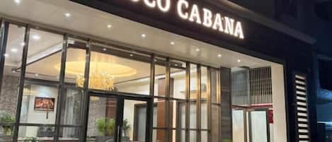 The exterior of the Coco Cabana hotel with a modern facade, large glass windows, and a well-lit entrance.