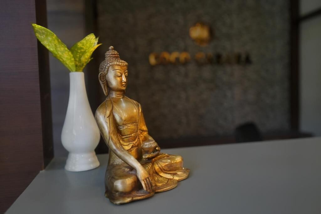 Kultainen Buddha-patsas pöydällä, valkoisen maljakon vieressä, jossa on vihreitä lehtiä.