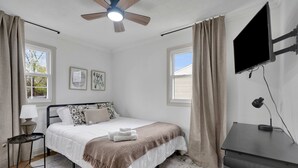 A bedroom with a bed, a desk, a ceiling fan, a TV, and two windows with curtains.