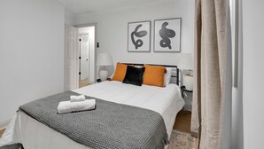 A neatly made bed with a gray blanket and a folded white towel, a bedside table with a lamp, and two abstract wall art pieces.