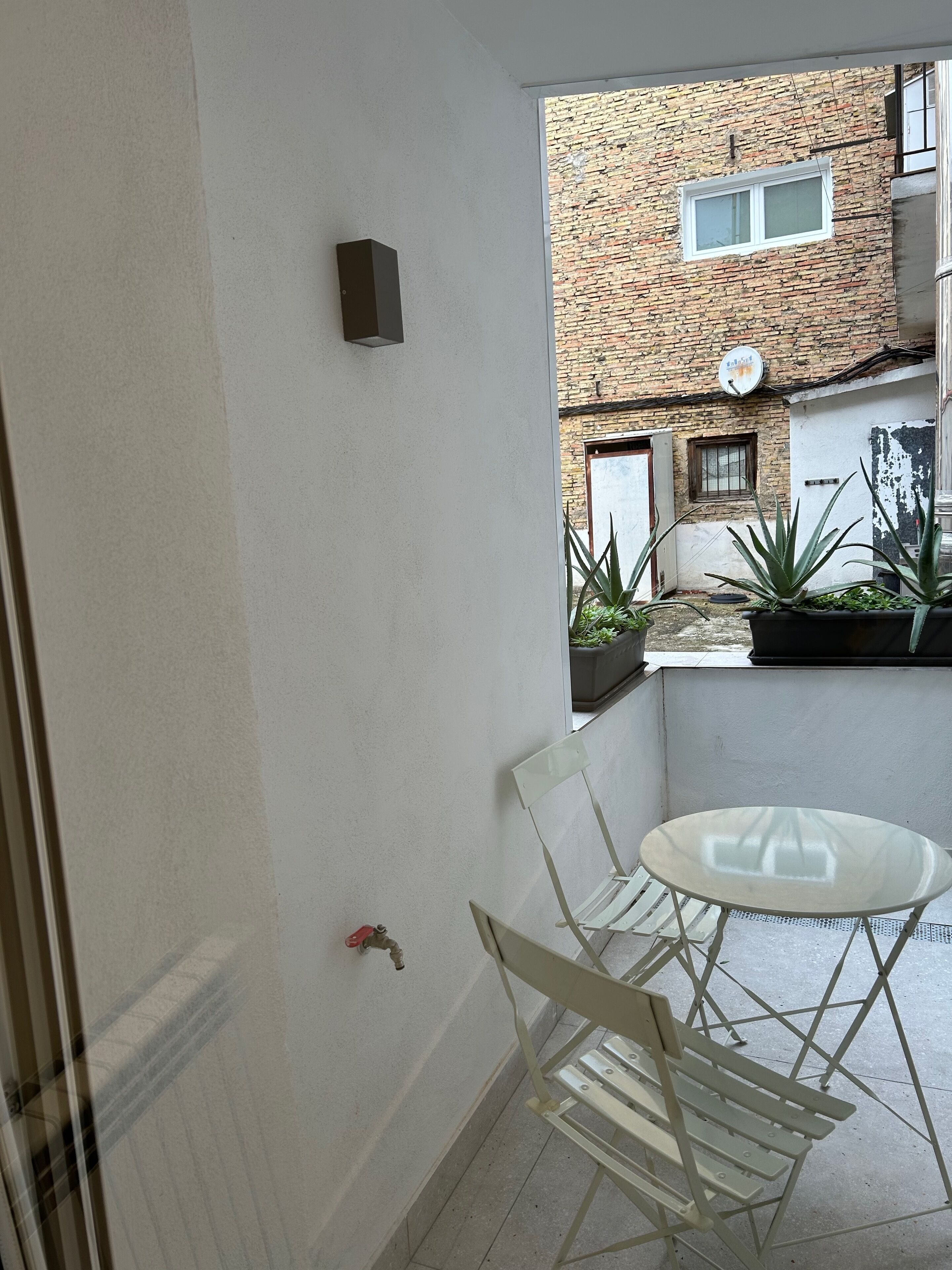 A balcony with a white table and chairs, a potted plant, and a brick building in the background.