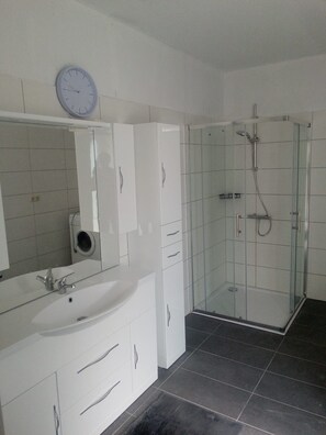 Une salle de bain moderne avec un lavabo blanc, un miroir et des meubles de rangement, une douche en verre et une horloge au mur.