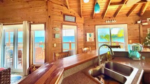 Ein Interieur einer Holzhütte mit Küchenbereich, einem Esstisch und Blick auf den Ozean durch große Fenster.