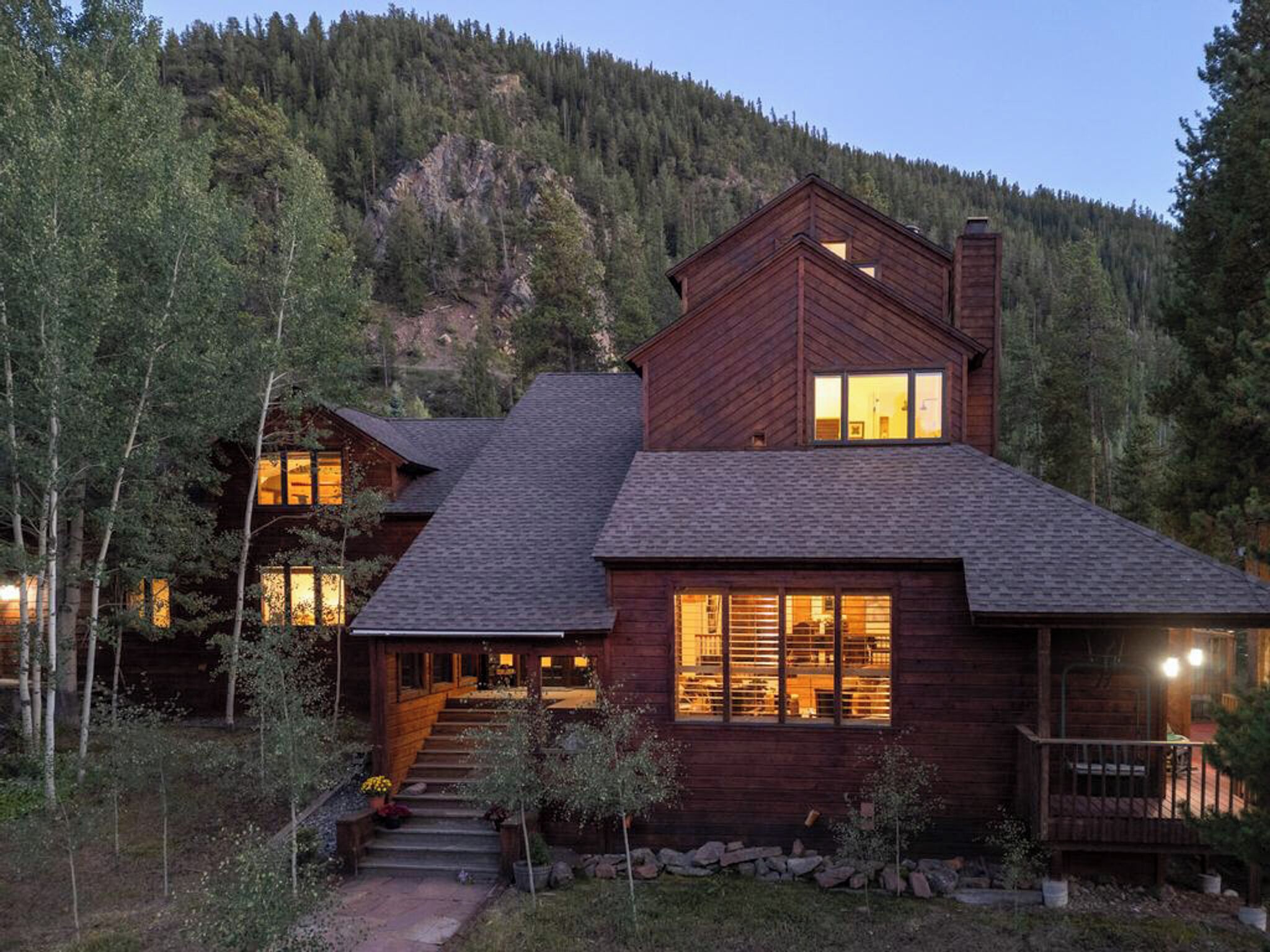 A large, multi-story wooden cabin with a steep roof, surrounded by trees and a mountain backdrop.
