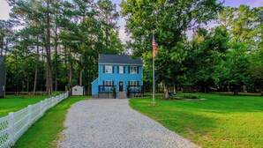 A two-story house with a blue exterior, a gravel driveway, and a white picket fence. - Premier Homestead Retreat on 1.5 Acres: Pets, Spa, Firepit, Chickens, King, Gym (Wake Forest)