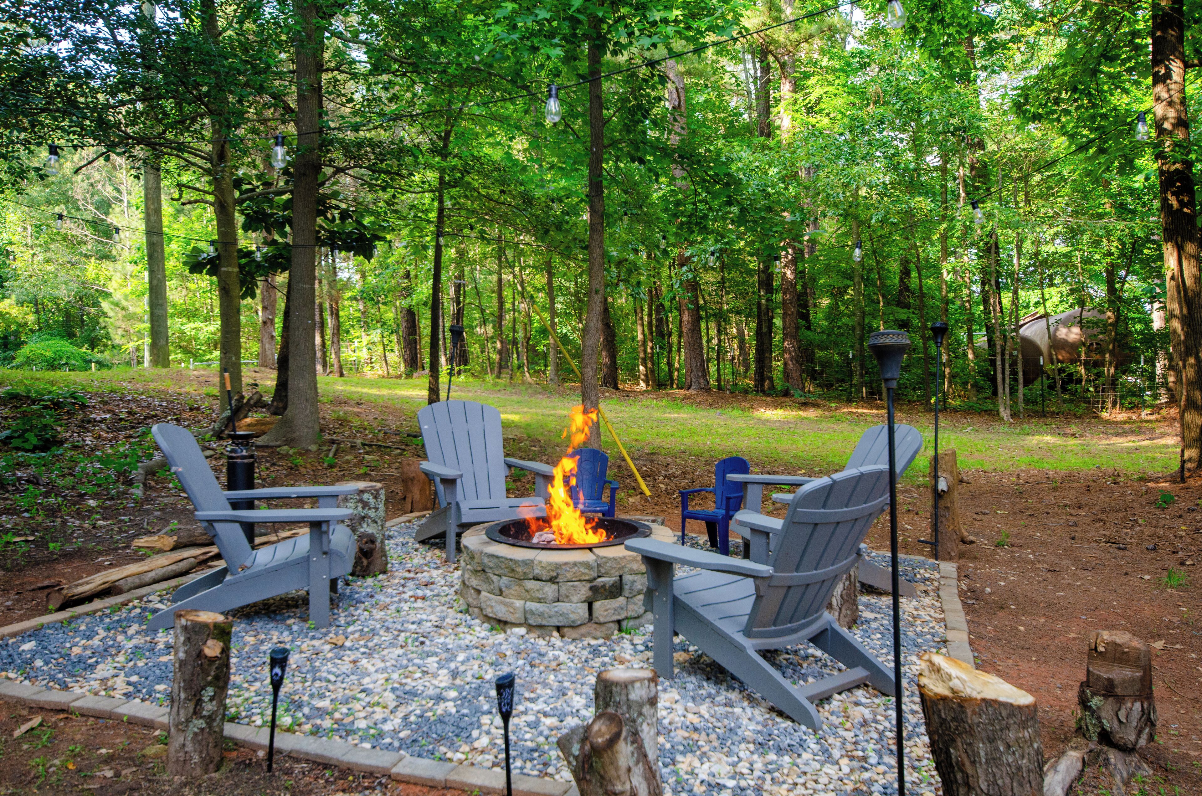A fire pit with four Adirondack chairs arranged around it, set in a forested area with trees and a grassy field.