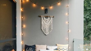A cozy balcony with a wooden bench, a cushioned seating area, string lights, and a macrame wall hanging. - Peace Family Vacation Home, Swimming Pool, Barbecue Grills and Gym (Playa del Carmen)