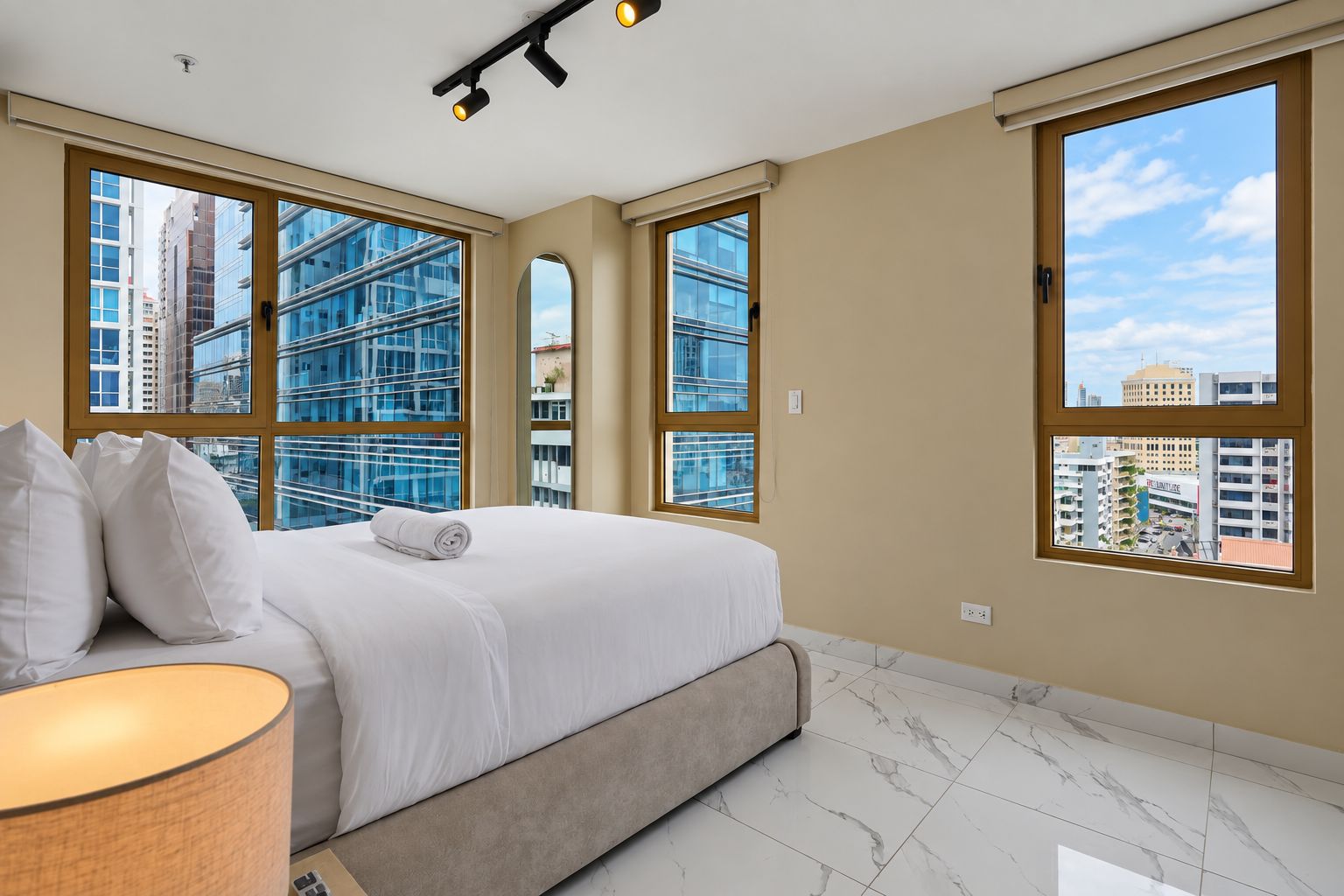A hotel room with a bed, a lamp, and large windows offering city views.